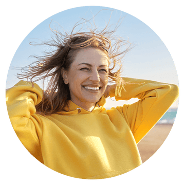 Smiling woman in a yellow hoodie enjoying the beach with wind in her hair, radiating joy and happiness.