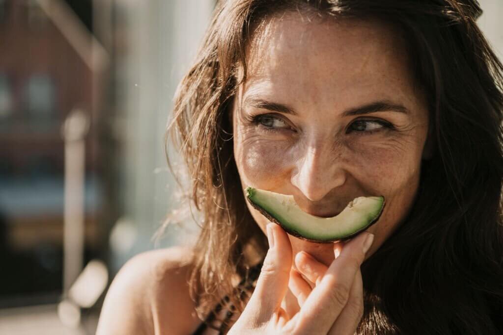 Woman playfully smiling with a slice of avocado in her mouth, showcasing a joyful moment outdoors.